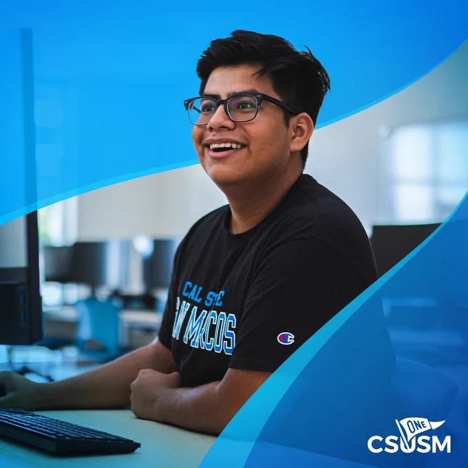 Student wearing a CAL state San Marcos T-shirt, sitting at a computer, smiling.