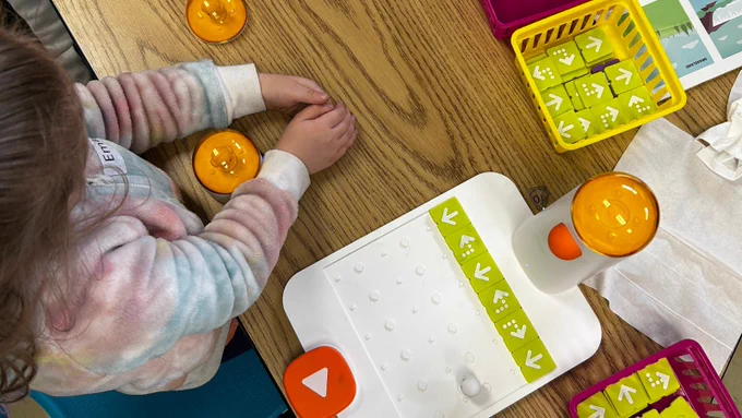 A top-view of a student creating patterns with the Matatalab platform. Green and purple tiles are in a basket nearby and the little robot sits in front of the student.