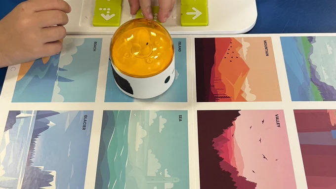 A close-up view of a the Matatalab robot on top of tiles picturing different landscape scenes. The pictured tiles are a beach, glacier, island, sea, mountain, and valley - all with soothing shades of blue and pink. A student's hand can be seen in the top of the image where they are arranging tiles on the Matatalab board. 