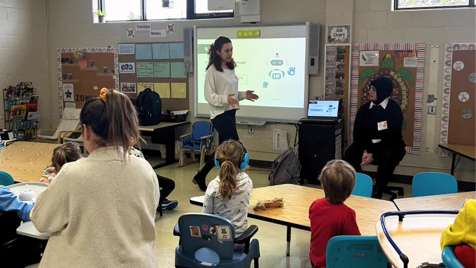 Two Engineering Outreach program instructors are at the front of the classroom with one standing in front of a projector gesturing with her hands and another sitting in a chair. Children are sitting on blue chairs at short brown desks and attentively listening to the instructors. The classroom is colourful with photos, and learning material attached to bulletin boards on the walls.