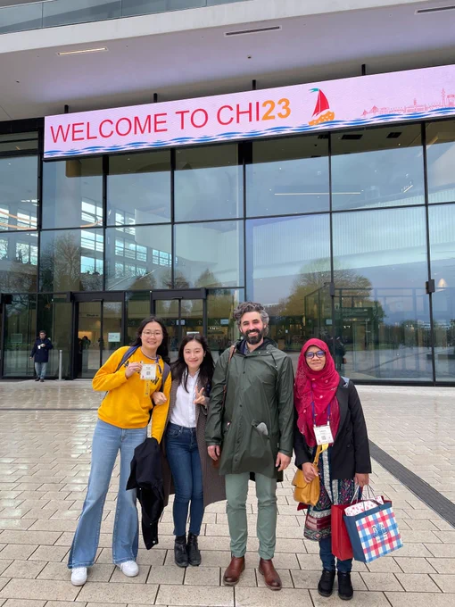 Four people standing in front of the Congress Center Hamburg.  "Welcome to CHI23" sign is in the background and above their heads.