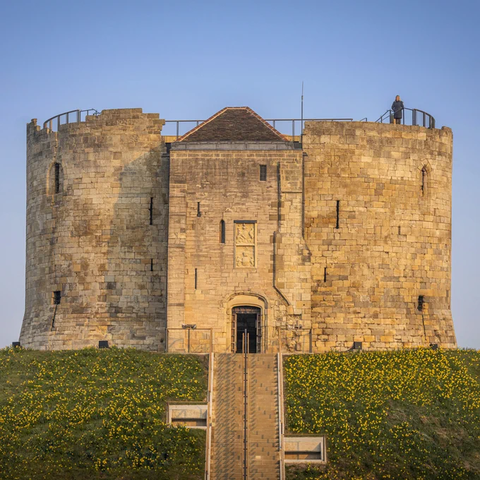  An image of a round tower atop a large man-made mound. The tower is constructed from light beige stone, with several thin slits for windows. Its original roof has been destroyed and been replaced by a new wooden roof and walkway. There are steps at the front of the mound leading up to the tower's arched doorway. 