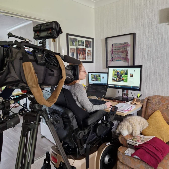 Hannah sitting in her powerchair in front of the computer being filmed by a camera behind.