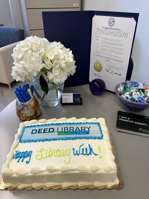 Photo that contains a cake with the words DEED Library and Happy Library Week! The photo also contains the signed proclamation from Governor Walz declaring this week as Capitol Area Library Consortium Week, a bouquet of flowers and assorted candies in a dish. 