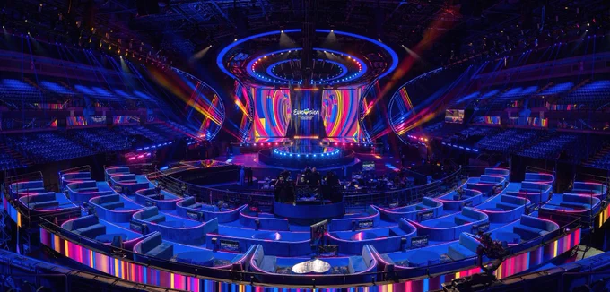 The Eurovision Song Contest 2023 stage is bathed in blue, pink and yellow strips of light with delegate seating areas facing it in a semi circle. The stage features a rectangular portion, with a catwalk jutting out from the centre to a circular stage, ringed by lights.