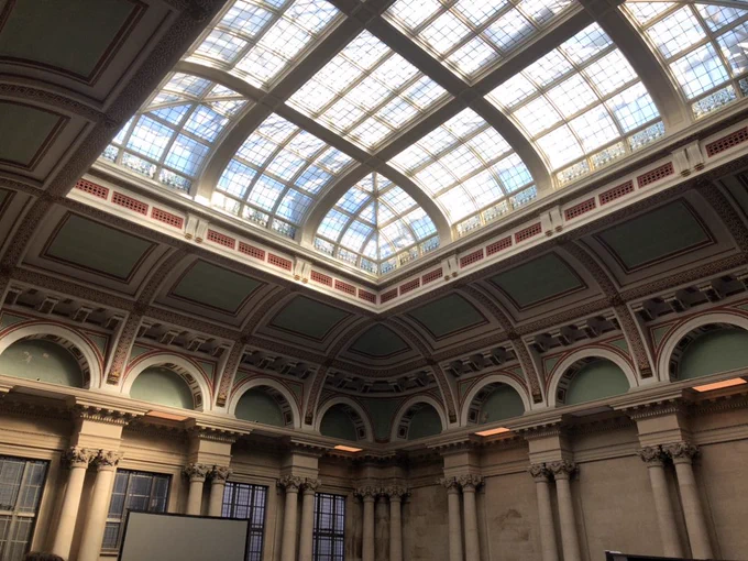 Photo of the conference venue, which looks like it could be a ballroom or spa - a big square room with stone walls with decorative columns around the walls. The top of the walls curve into the ceiling, and are painted green, red and cream. The ceiling is a huge domed window taking up most of the room. There’s a row of windows at the back of the room, and a projector screen set up in front of them.