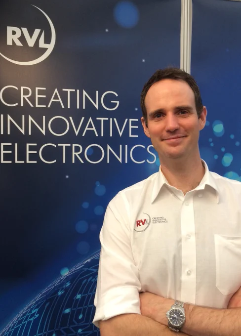 Portrait photo of Peter Hall, Software Director, smiling and looking ready to answer questions about the firm of electronic design engineers that he is employed by. He is standing in front of a blue background with white lettering that say "Creating Innovative Electronics", and the RVL company logo is above these words. Peter is wearing a white shirt which also has the RVL company logo on it, on the front left.