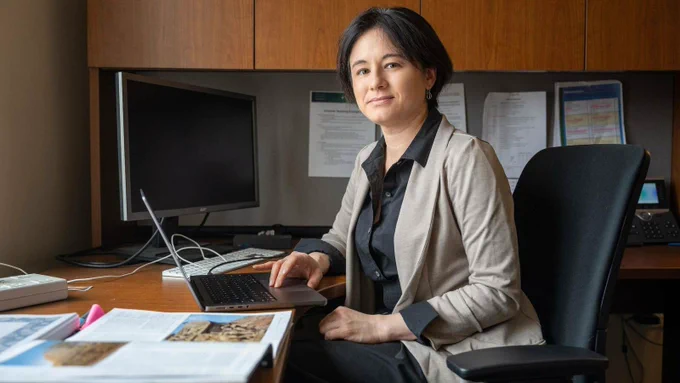 Mari Yoko Hara, a Japanese-American woman sits at a desk wearing a khaki sweater over a black button down.