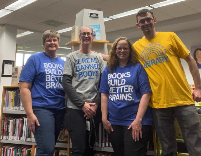 Library staff members standing in front of our new book shelves, wearing RayGun t-shirts that say, Books Build Better Brains, I Read Banned Books and Everyone Is Welcome at the Library.
