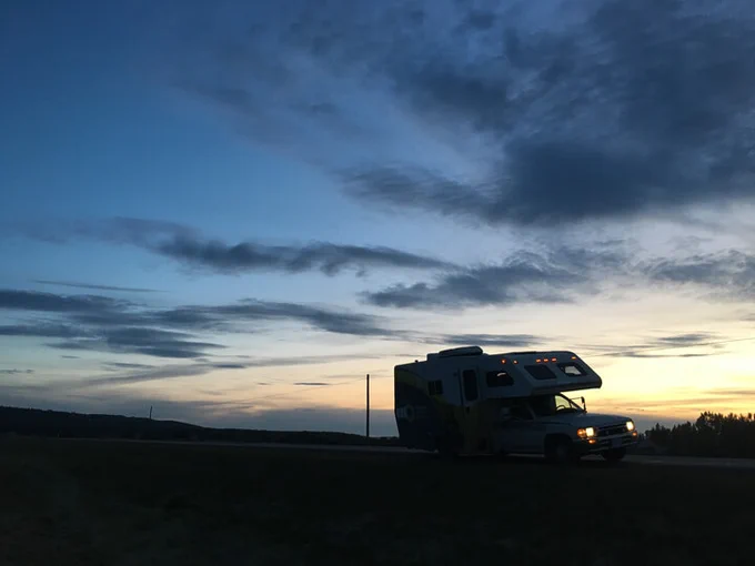 Image is of an RV being driven on a road around dusk time. The sky is blue and yellow with clouds in it.