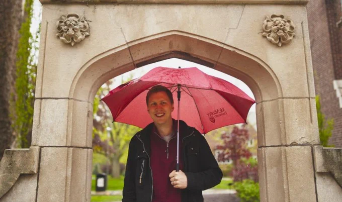 Drew Higgins stands under the Edward's Arch holding an open McMaster umbrella above his head.