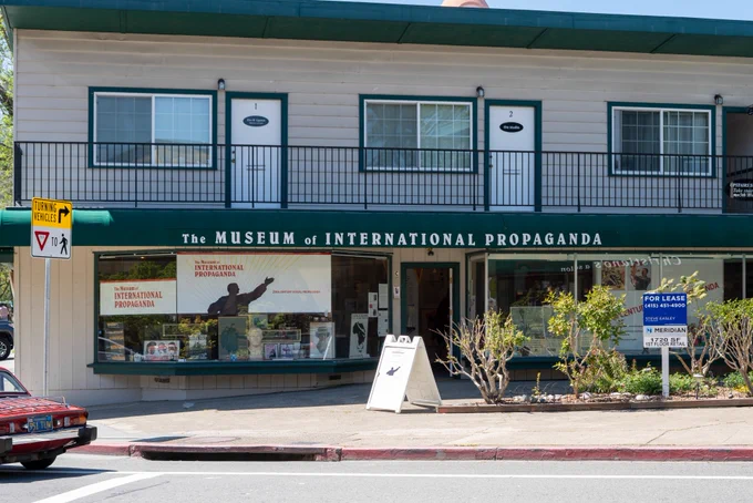 Photograph of a storefront on a street corner. A sign above the storefront reads “MUSEUM OF INTERNATIONAL PROPAGANDA.” A small sign in front of the storefront reads “WE ARE OPEN.” Posters of propaganda art are visible through the storefront windows.
