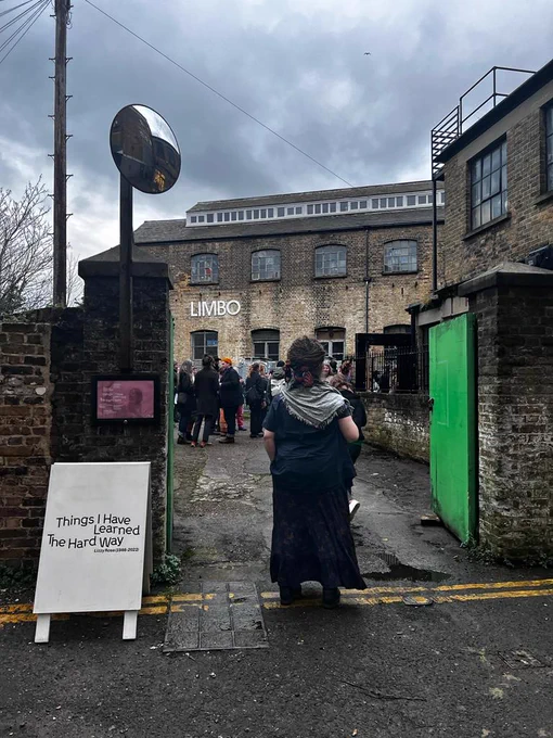 The exhibition venue limbo. A brick building with a LIMBO sign on the front. People gather round in the courtyard for Lizzy's Private Viewing. There is a white aframe with the promotional branding of Things I Have Learned The Hard Way on the front. 
