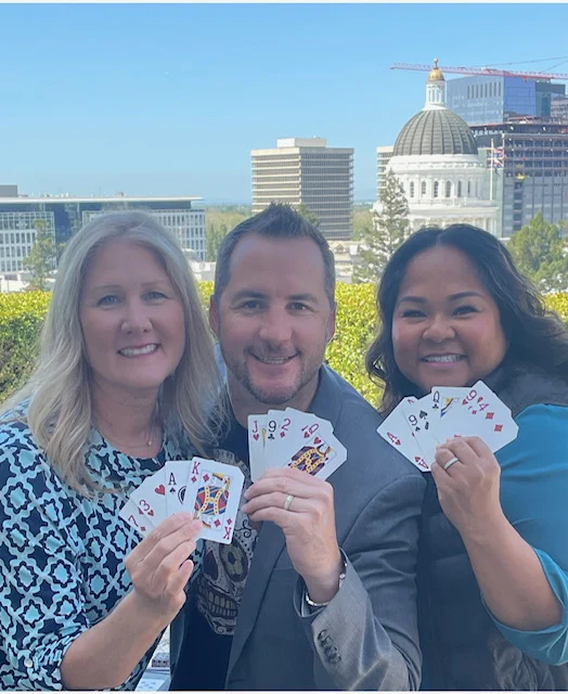 Three people holding playing cards on a patio with views of the California State Capitol.