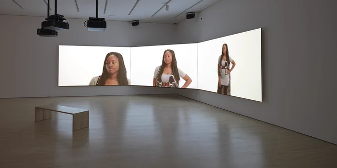 Picture of 3 video screens on corner walls of an empty gallery with images of a person on each screen
