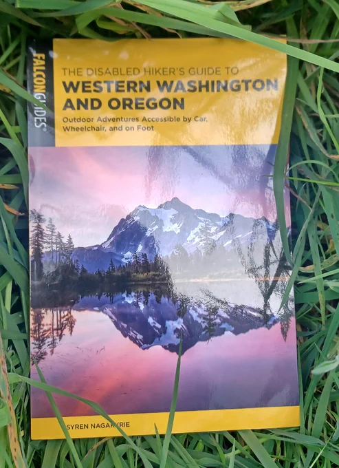 A paperback book with a yellow cover and black lettering saying "Falcon Guides - The Disabled Hiker's Guide to Western Washington and Oregon: Outdoor Adventures Accessible by Car, Wheelchair, and on Foot - Syren Nagakyrie." The center of the cover has a picture of a lake reflecting the snow-covered mountains, trees, and bright pink and blue sunset in the background.