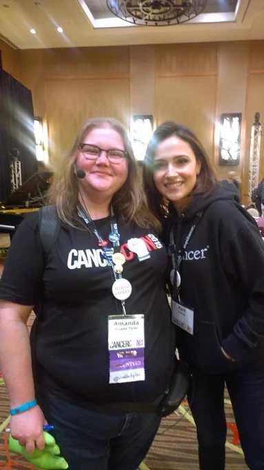 Me, smiling in a black CancerCon shirt, button-layden name tag, and ChatterVox microphone set standing next to Italia Ricci, also smiling in a black Stupid Cancer hoodie.