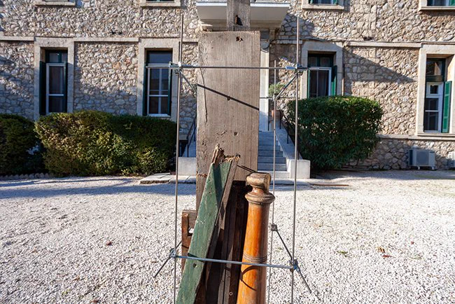 A sculpture made of found wooden materials:  a shelf, a stake painted green, a table leg , helf loosely together by metal rods. It is sunny. the sculpture is placed at the entrance of a thick walled stone building. The artwork is by previous resident Elin karlsson and is placed outside the british School at Rome builsing.
