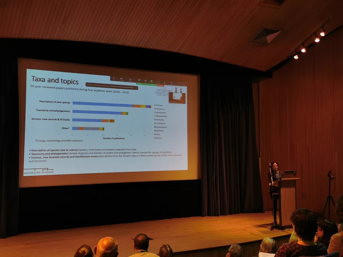 View towards a stage in a large lecture theatre. A speaker stands behind the podium and a huge screen displays a slide with a graph of the number of publications from Manchester Museums invertebrate collection