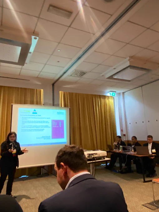 IARIA Special Track Digital Accessibility Rednerin Prof. Möhring (THM) vor einer Präsentation des lila Sammelband Covers Menschen im Publikum und am Tisch mit Laptop und dem THM-Stand zu  Barrierefreie Info-Materialen und Medien.