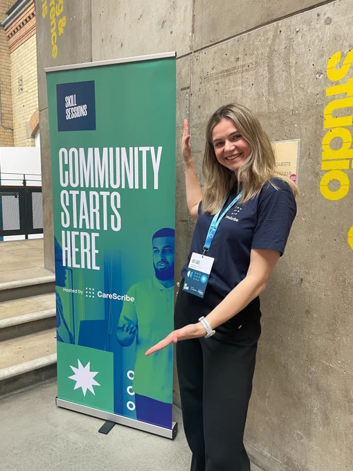 A photo of Lucy who is standing next to a Skill Sessions stand with her arms pointing towards the stand, smiling.