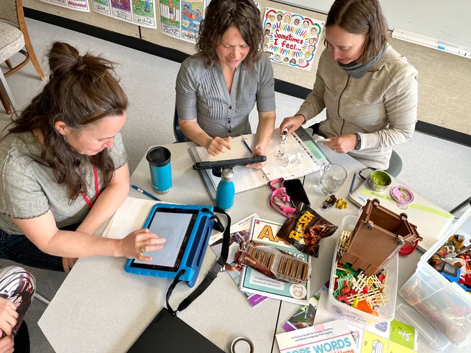 Three women sit  at a table with crafting materials and iPads.