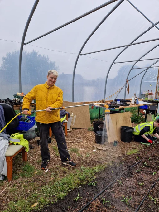 Andy, a white man in a yellow coat, looks exceptionally happy whilst holding a hose and watering the soil.
