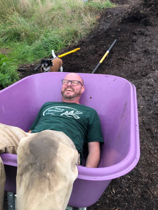 Jon, a white man wearing glasses and a green t shirt, lies laughing in a wheel barrow. Banter.