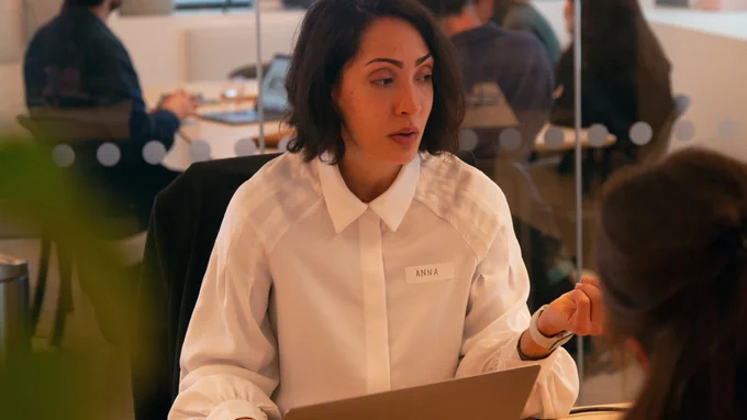 One of our portfolio reviewers, Anna, sat at a desk with a laptop, reviewing a portfolio and giving feedback. Behind her in another meeting room, Ed is giving feedback to another group of attendees. 