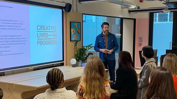 Our creative director, and one of our portfolio reviewers, Ed standing in our studio presenting our intro deck about the event to our attendees. The attendees are sitting on stools and chairs and taking in Ed’s presentation. 