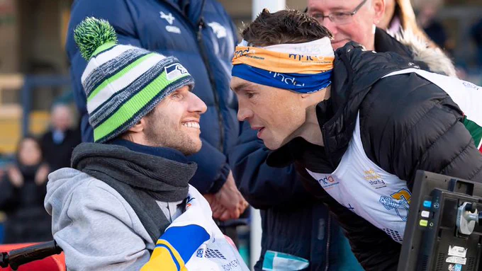 A photograph of Rob Burrow and Kevin Sinfield. Rob is smiling looking at his friend Kevin, who is leaning over Rob's wheelchair to talk closely to him. Both rugby players are wearing charity fundraising vests and are surrounded by a group of people at a fundraising event. 