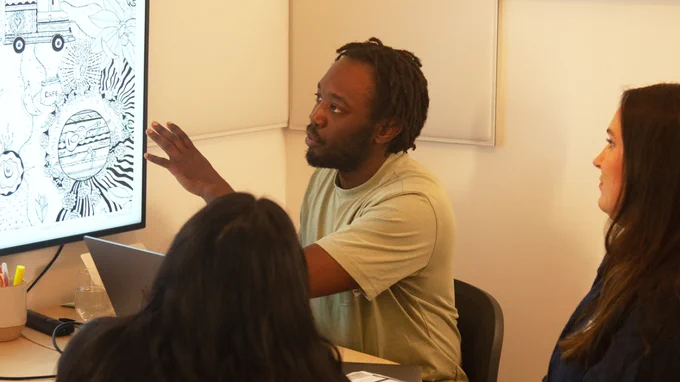 Our portfolio reviewer Marc, sad in a meeting room with three attendees. He’s gesturing to a large screen which is showing work from a portfolio, and is discussing it with the attendees.  