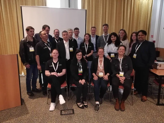 18 Personen in einem Konferenzraum, lächeln in die Kamera.