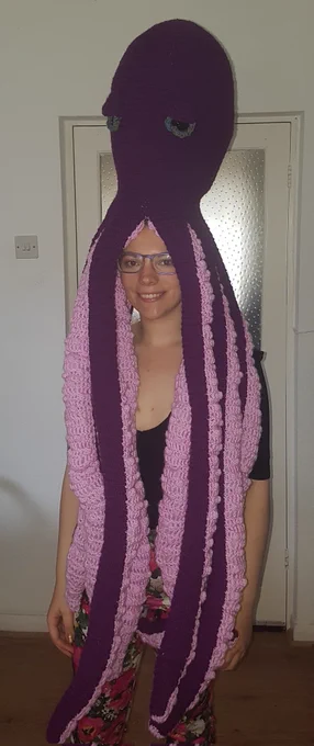 Photo shows Claire, a white woman with purple glasses, with a large purple crochet octopus on her head. The octopus head is around 2 ft tall, and the tentacles extend down to Claire's knees.
