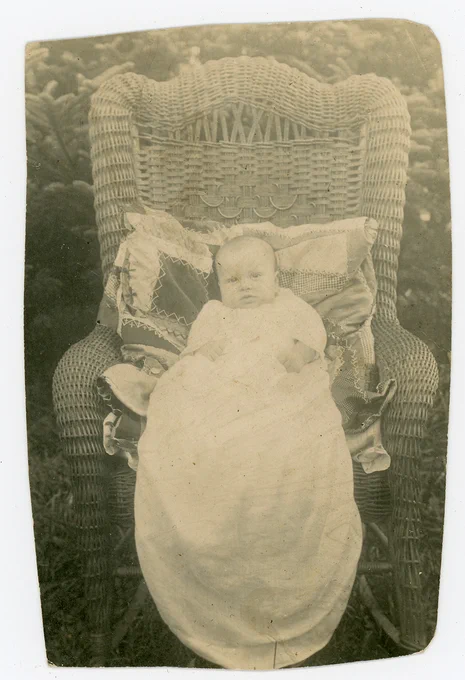 A baby in a long white garment is pictured lying on pillows in a wicker chair.