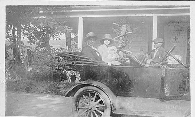 Four people are seated in an automobile parked in front of a house. One is a baby in a bonnet. The automobile and clothing indicate that the photograph was probably taken in the years around 1920.