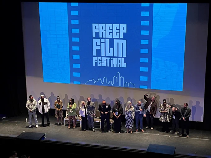 A lineup of people involved in the making of the film Coldwater Kitchen and the Freep Film Festival standing on a stage against a screen with the words: Freep Film Festival