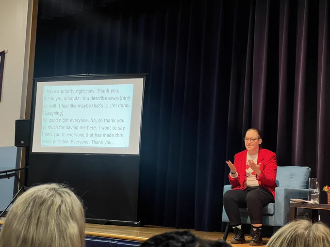 Author Sara Nović, on a chair on stage, discusses her book, True Biz, next to a transcription board.