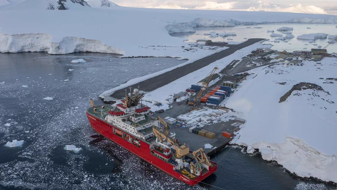 An aerial photo of the SDA ship docked in the icy waters at Rothera. the dock and the runway are dark grey against an otherwise white landscape.