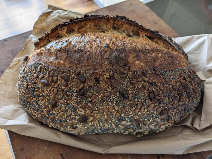 A lovely fresh, seeded loaf on a brown paper bad on top of a wooden chopping board 
