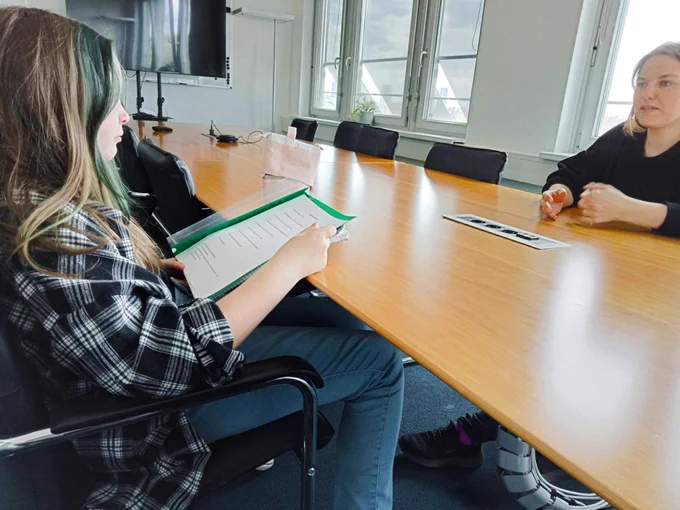 Mila and Judith are having a conversation in the conference room. Mila sits on the left side, a folder with questions in her hands. Judith sits opposite her and answers her questions. There are more chairs at the long table and a large screen at the end of the room.