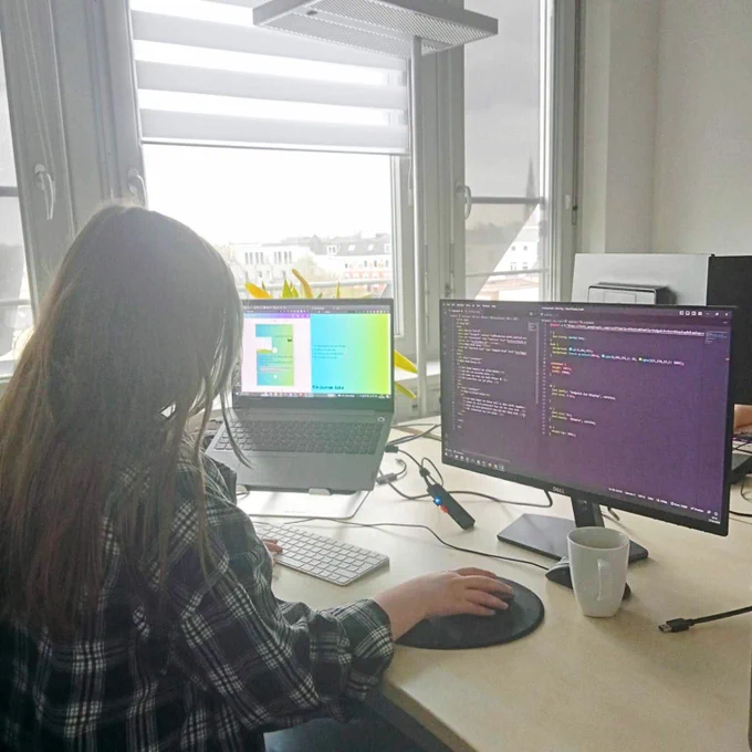 Mila is sitting at a desk working on the computer in VSCode on the external screen, one hand on the mouse, the other on the keyboard. On her left is a laptop with the current state of the website she has created and the draft of it. 
In the background is a window with a view of the rooftops of Hamburg.