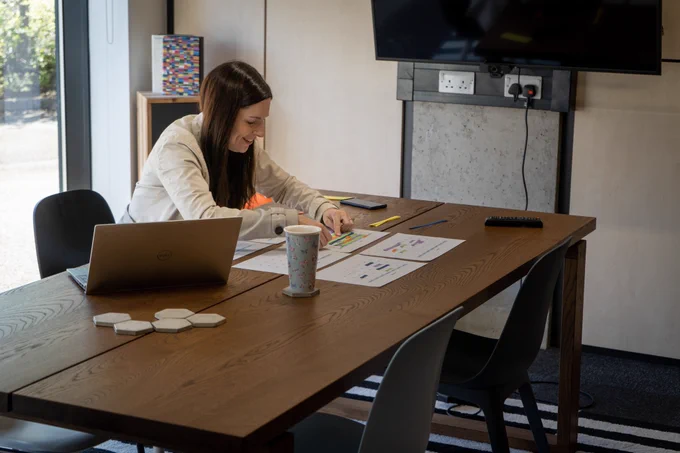 Zoe from Team Epix is sat at a wooden table with pieces of paper laid out in front of her, planning projects the old fashioned way with coloured strips of paper to block out dates on the schedule. Also on the table is a laptop and a cup of coffee.