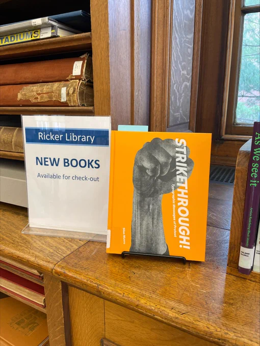 An image of a book titled "Strikethrough!" on display. The book is propped up on a wooden countertop; to the left of the book is a small sign reading "Ricker Library: New Books, Available for check-out". Horizontal folio bookshelves can be seen behind the sign. 