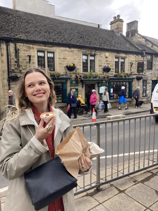 A woman is holding a Bakewell tart in front of the bakewell pudding shop in the centre of the town of Bakewell. She looks very happy.