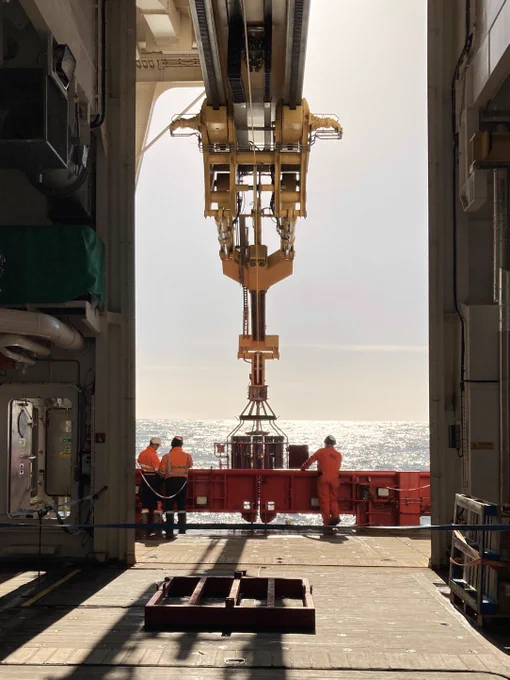 A long profile photo of the CTD on a yellow crane, taken from inside the dock area of the SDA. Scientists stand in profile against a shining Antarctic sea.