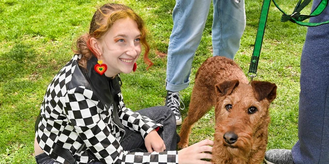 A student sits on the ground, smiling as they pet a rust-colored, floppy eared emotional support dog.