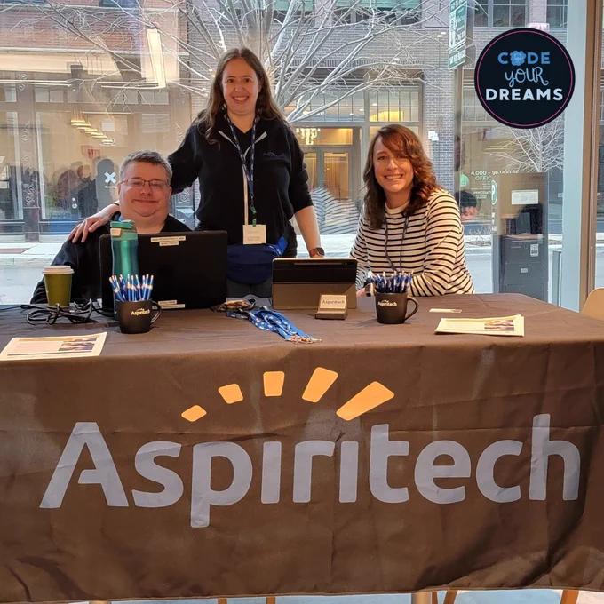 3 people posing in front of an Aspiritech table with laptops and Aspiritech branded items