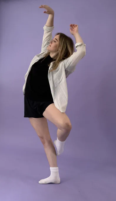 Female dancer is standing on one leg with a her other  leg raised and bent. Her arms are stretched above her head.