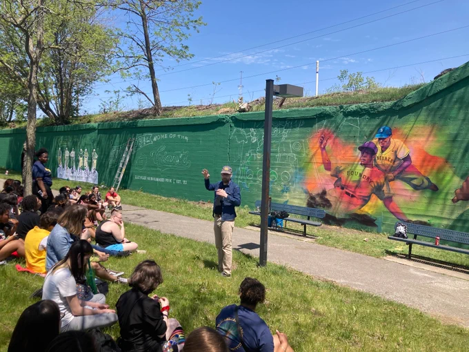 Man in front of mural wall speaking to group of students